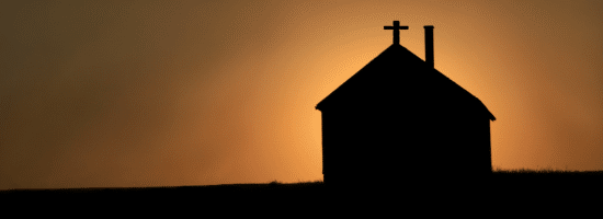 Silhouette of a small church with a cross on the roof at sunset, set against a gradient orange and brown sky.