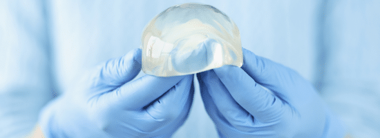 Gloved hands holding a transparent silicone breast implant against a light blue background.