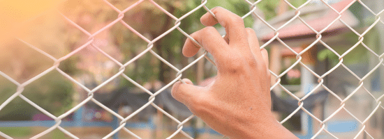 A hand grips a metal chain-link fence with buildings and greenery blurred in the background.