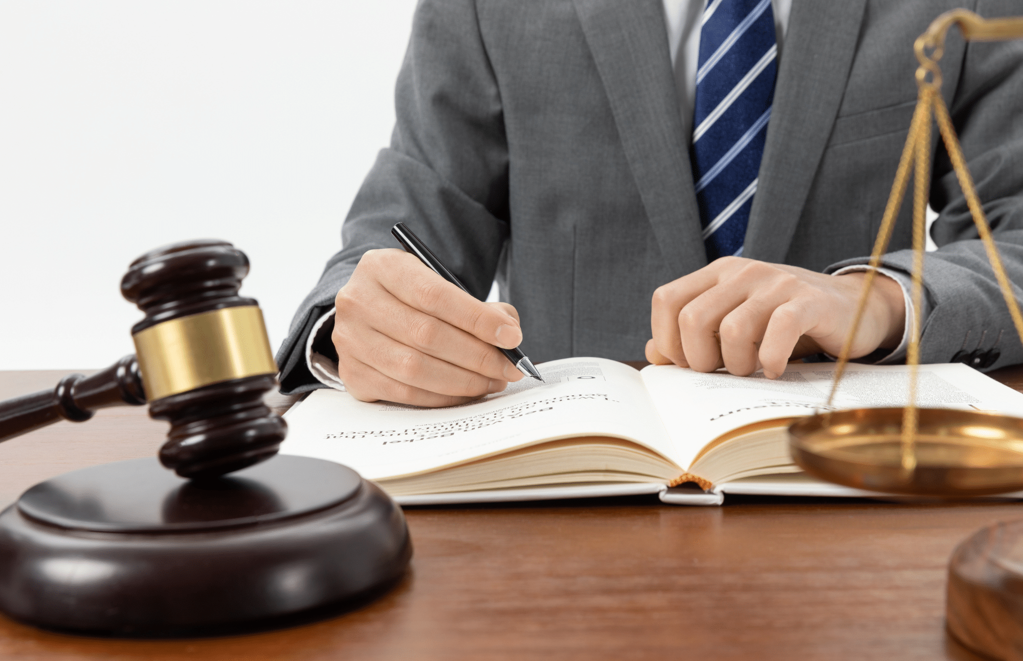A person in a suit writes in an open book at a desk with a gavel and scales of justice.