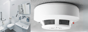 A white accessible bathroom with grab bars, a wall-mounted sink, and a mirror is shown on the left; a close-up of a white smoke detector on the ceiling is shown on the right.