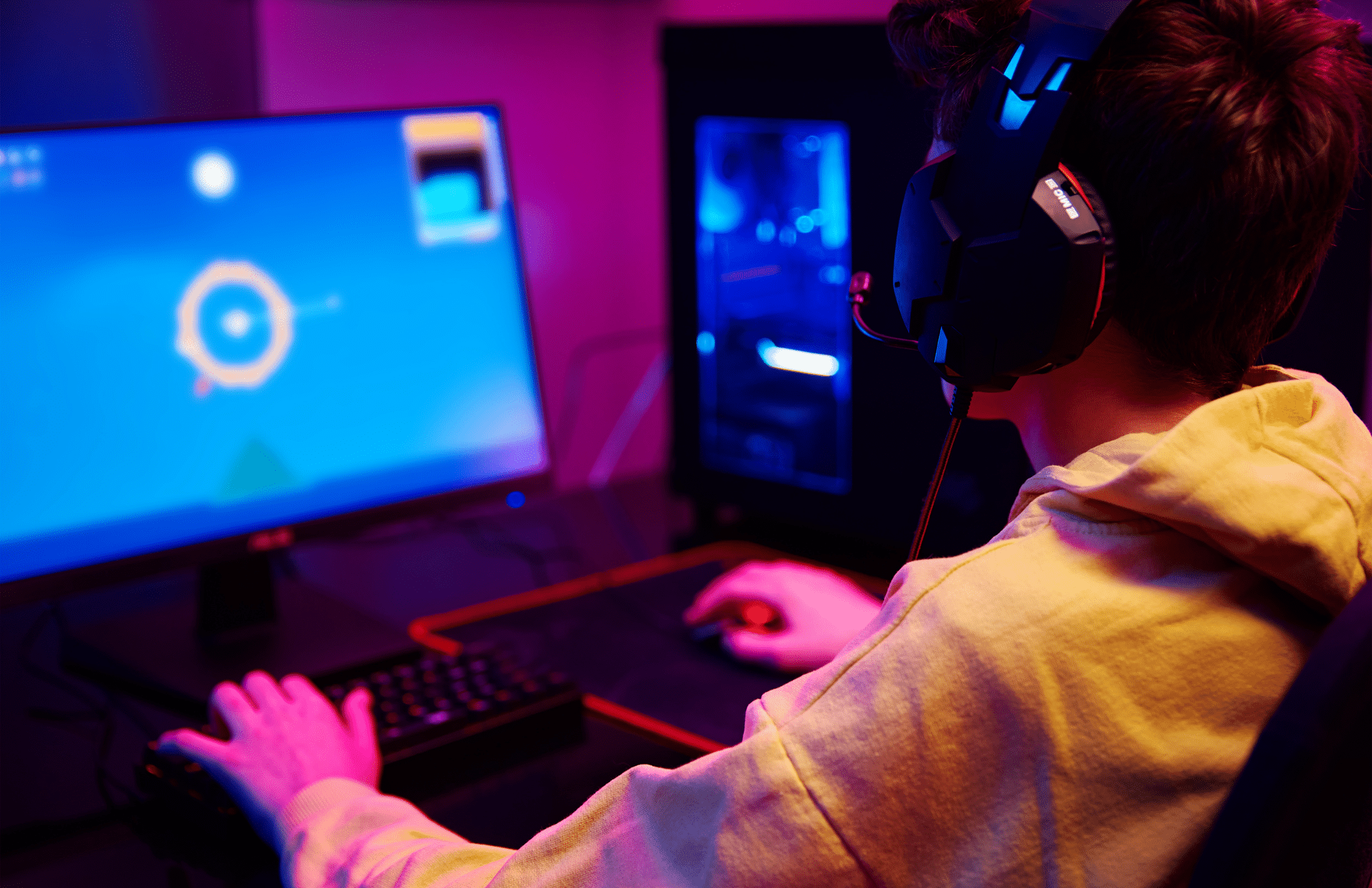 A person wearing a headset and yellow hoodie is playing a video game on a desktop computer in a room with colorful lighting.
