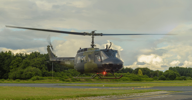 A military helicopter hovers low above a runway, with trees and cloudy sky in the background.