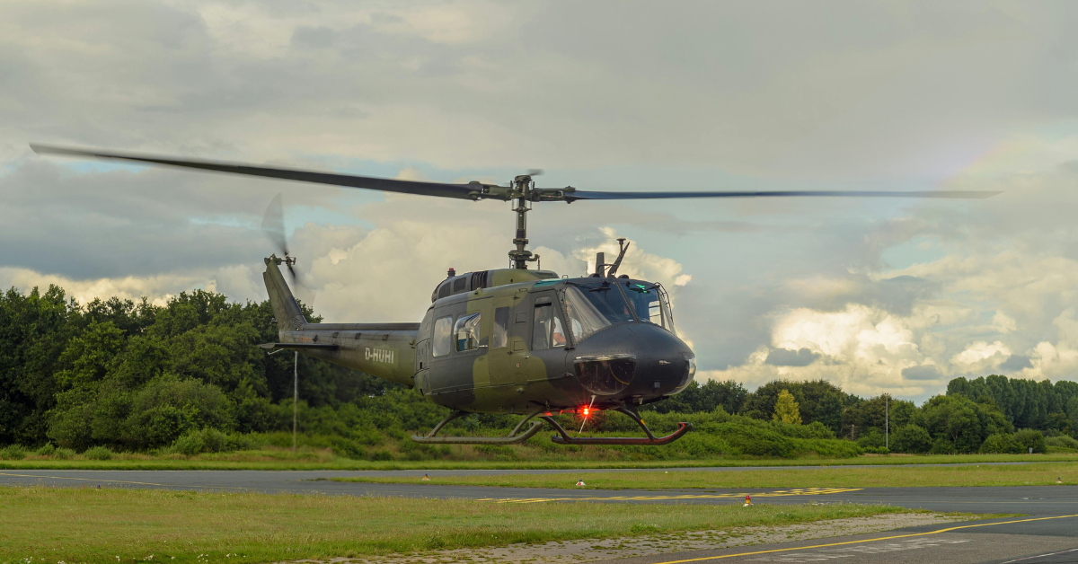 A military helicopter hovers low above a runway, with trees and cloudy sky in the background.