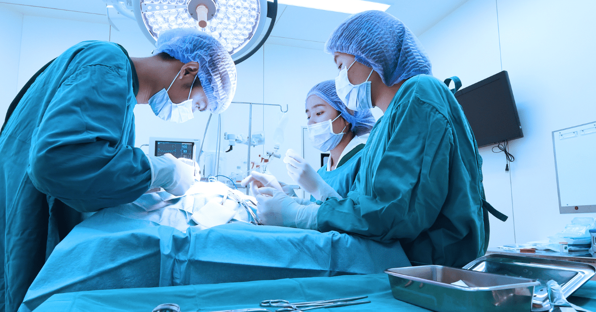 Three surgeons wearing blue scrubs, masks, and caps perform surgery on a patient in a brightly lit operating room with medical equipment visible.
