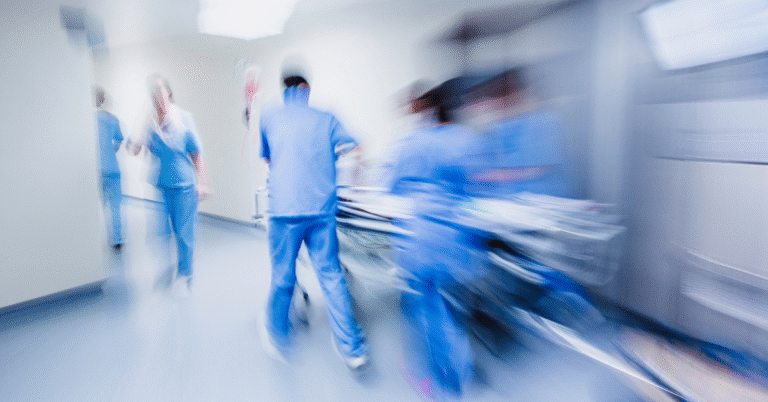 Blurred image of medical staff in blue scrubs moving quickly in a hospital corridor, possibly during an emergency situation.