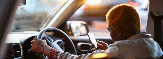 Person sitting in the driver’s seat of a car, holding the steering wheel with one hand and looking at a smartphone with the other hand.