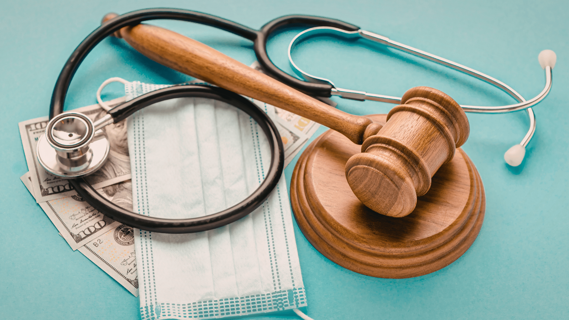 A gavel, stethoscope, face mask, and U.S. dollar bills arranged on a blue surface, representing themes of healthcare and law.