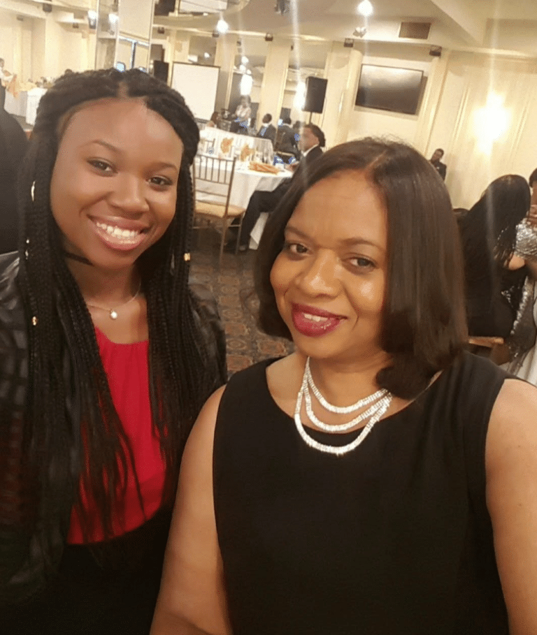 Two women pose and smile for a photo at an indoor event, dressed in semi-formal attire with tables and other guests visible in the background.