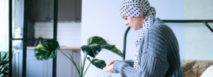 A person wearing a polka-dot headscarf and gray sweater sits indoors, looking down at something in their hands, with houseplants and a kitchen in the background.