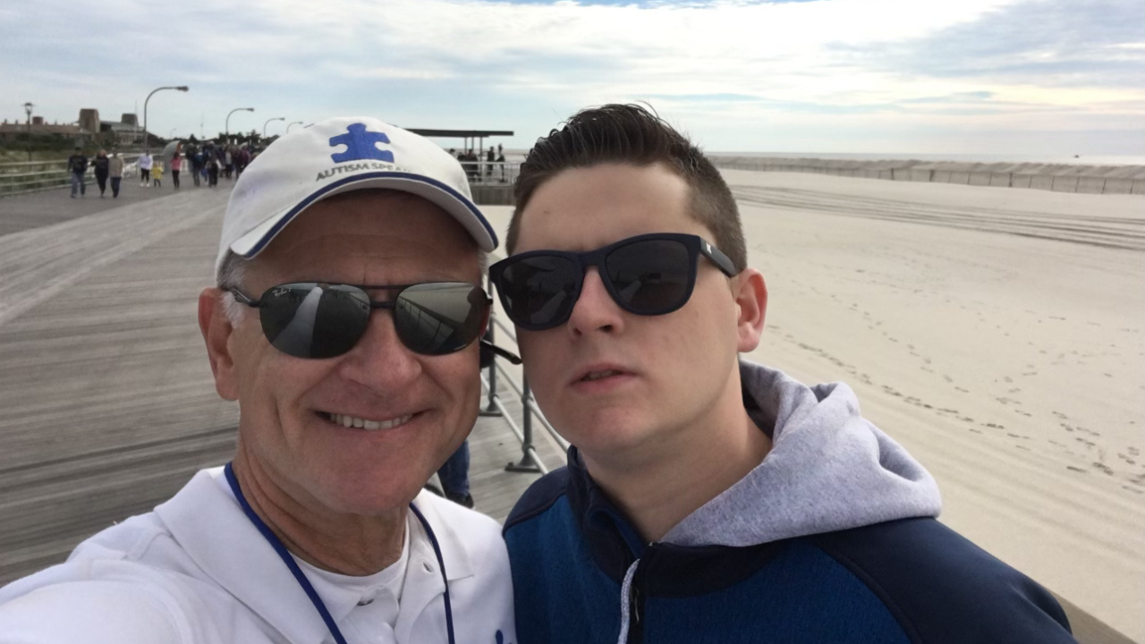 Two men wearing sunglasses pose for a selfie on a boardwalk next to a sandy beach on a cloudy day. One man is smiling; the other has a neutral expression.