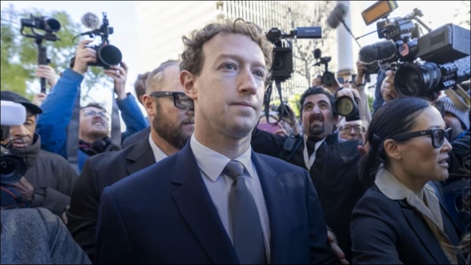 A man in a suit walks through a crowd of photographers and journalists, with cameras pointed toward him in an outdoor setting.