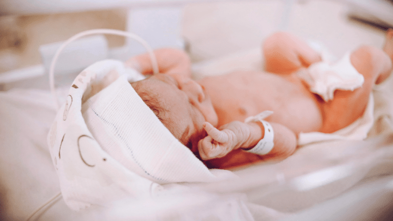 A premature newborn lies in an incubator, wearing a diaper, identification band, and a soft fabric cap with a monitoring wire attached.