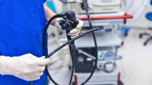 A medical professional in blue scrubs and gloves holds an endoscope in a clinical setting with medical equipment in the background.