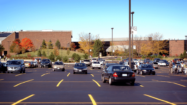 A large parking lot with cars parked in rows in front of a brick building on a clear day, with some trees in the background showing autumn colors.