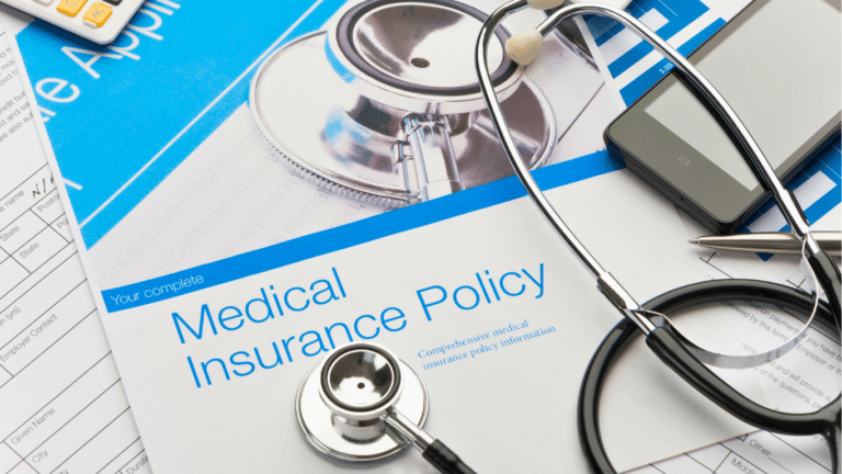 A stethoscope, smartphone, and pen rest on top of a medical insurance policy document and other paperwork.