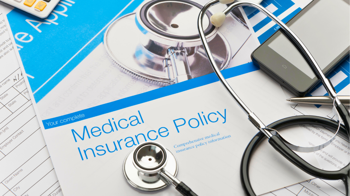 A stethoscope, smartphone, and pen rest on top of a medical insurance policy document and other paperwork.