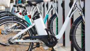 Row of bicycles parked at a bike-sharing station, secured in place, with black tires and white frames visible.