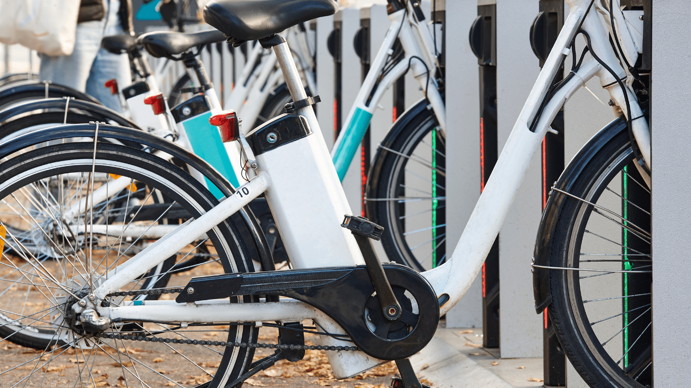 Row of bicycles parked at a bike-sharing station, secured in place, with black tires and white frames visible.