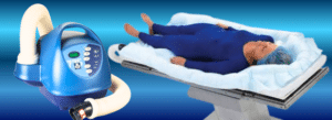 A medical warming device and hose are shown next to a mannequin lying on a padded hospital bed, covered with a blue suit and surrounded by a warming blanket.