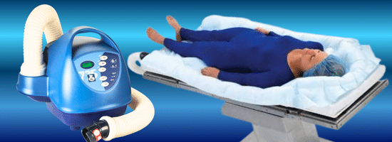 A medical warming device and hose are shown next to a mannequin lying on a padded hospital bed, covered with a blue suit and surrounded by a warming blanket.