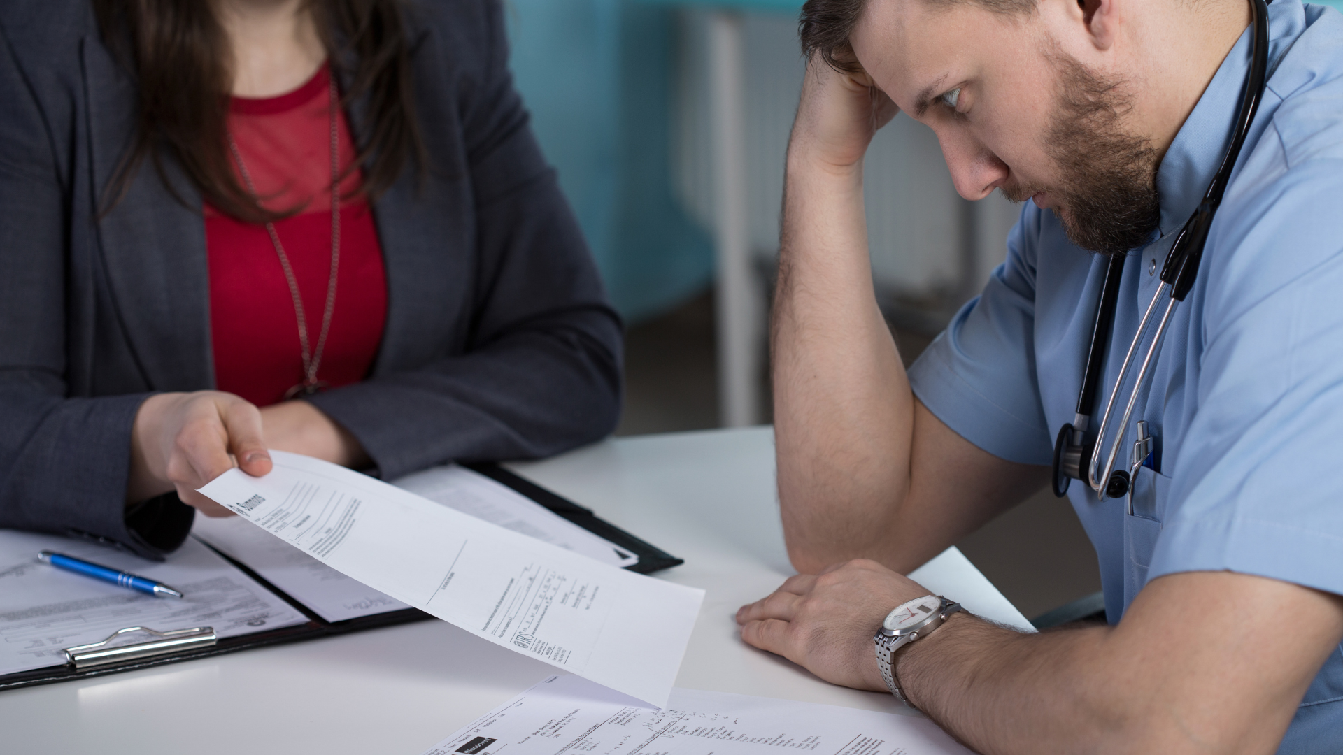 A doctor sits at a desk looking stressed while a woman in business attire shows him paperwork; forms and a clipboard are on the table.